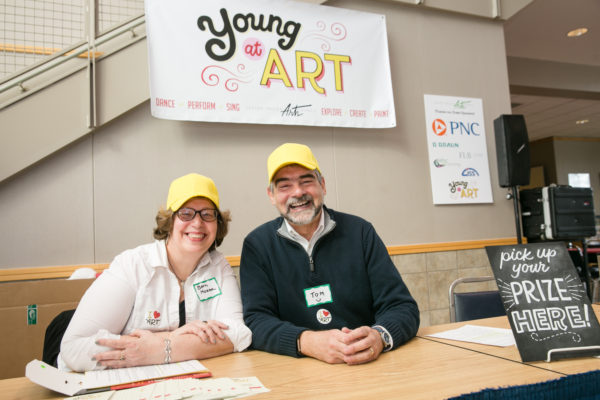 Two people wearing yellow baseball caps and smiling at the camera sit at an event sign-in table. In the background and on the table are colorful signs and posters for the event.