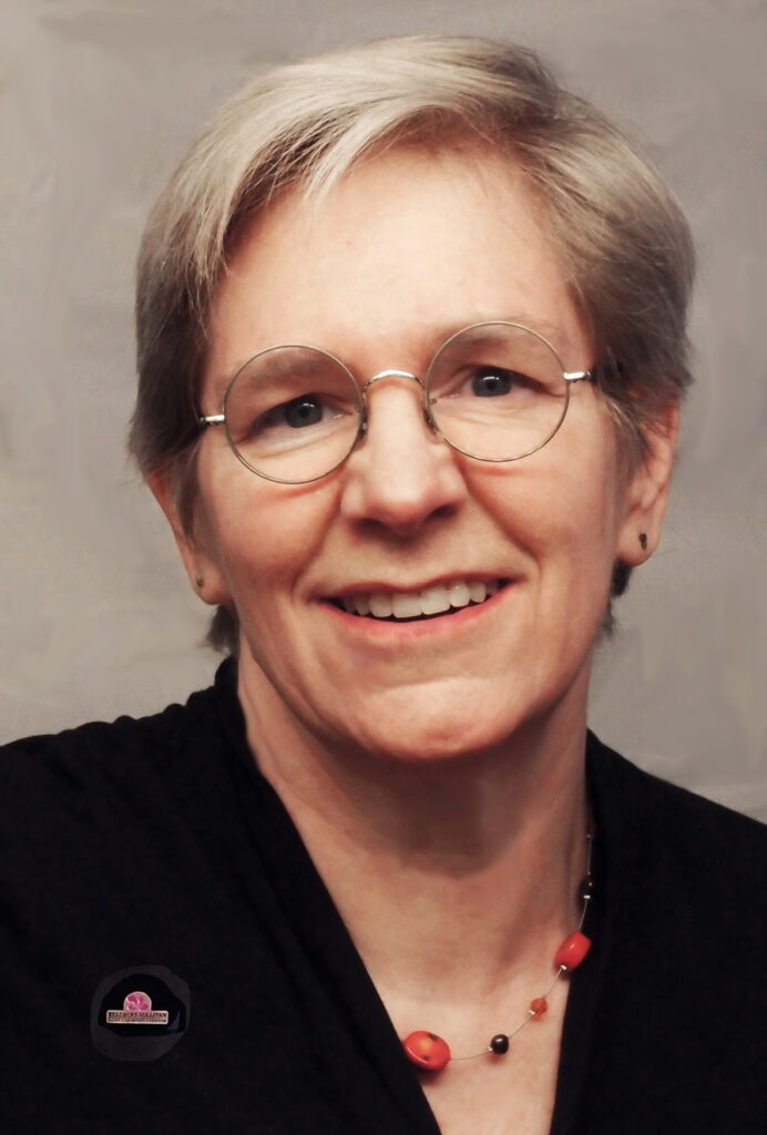 Liz, a white woman with short hair and glasses, is shown from the shoulders up looking at the camera and smiling