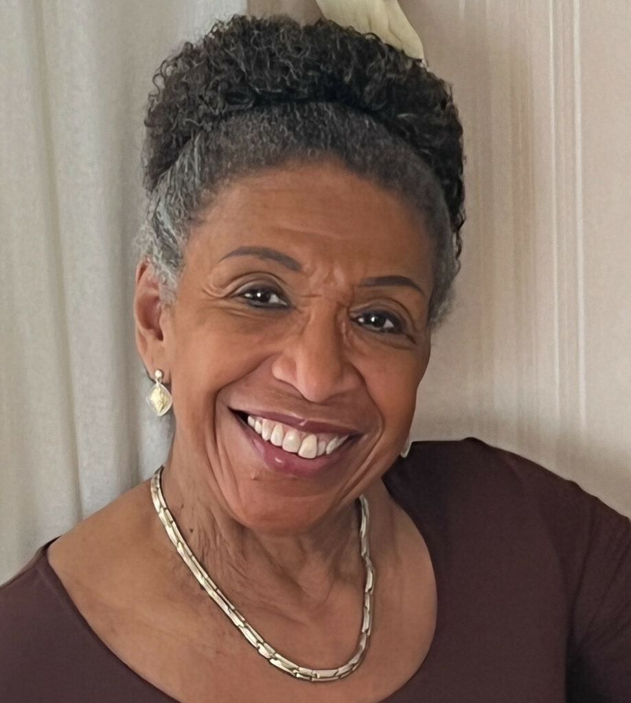 A photo of Bronwen Andrea Okwesa, a Black woman with dark hair worn in a bun on top of her head and smiling at the viewer. She is wearing earrings, a necklace, and a dark red top.
