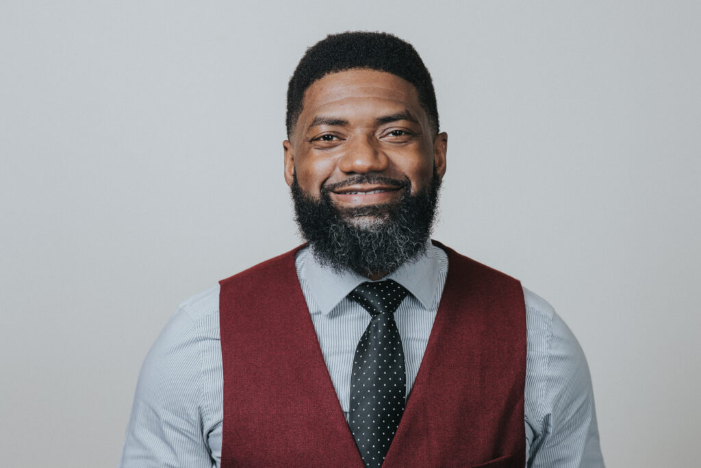 Tyrone Russell, a Black man with short black hair and a beard, wearing a dress shirt with a tie and a red vest. He is looking directly at the camera and smiling.