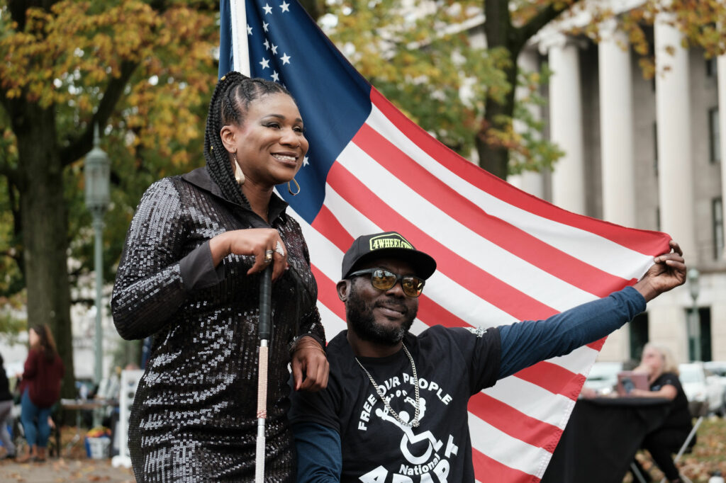 Two figures side-by-side. On the right, the seated figure extends his left hand to hold the corner of an American flag. Next to them, another figuring wearing a black, sparkly dress, smiles.